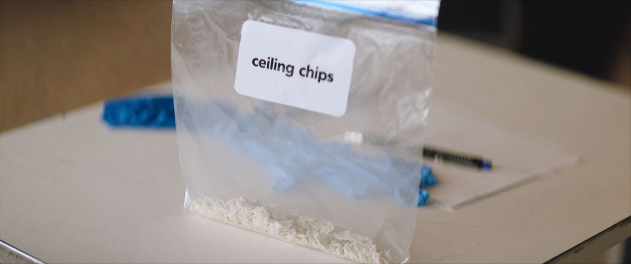 Clear lab sample bag holding chips of ceiling texture beside nitrile gloves on a work table