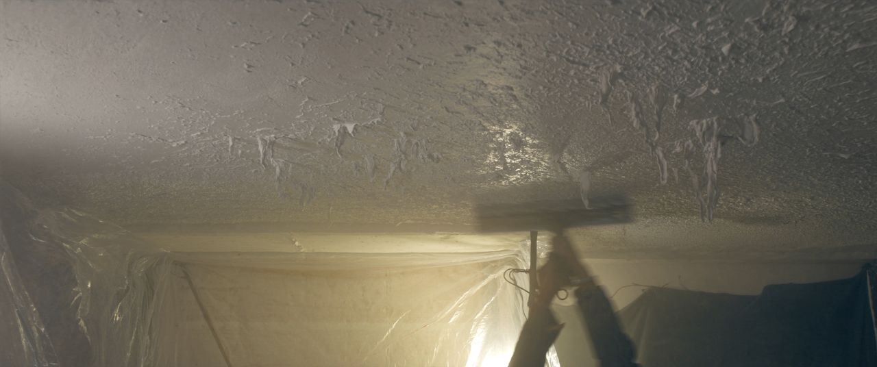 Roller soaking popcorn ceiling texture inside a plastic-contained room